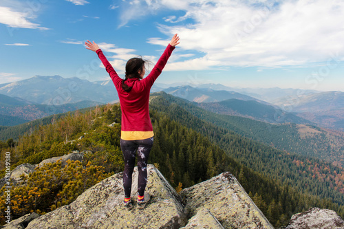 The young girl at the top of the mountain raised her hands up on blue sky background. The woman climbed to the top and enjoyed her success. Back view