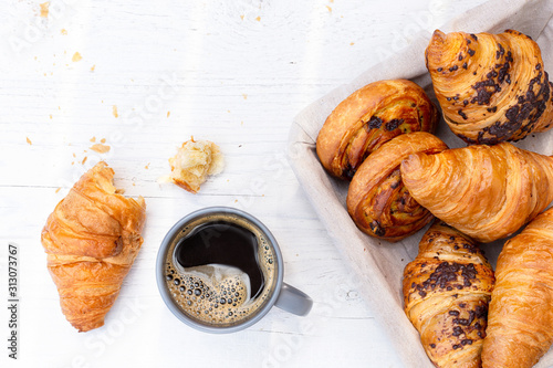 Continental breakfast with black coffee and basket of pastries. Half eaten on white wood from above.