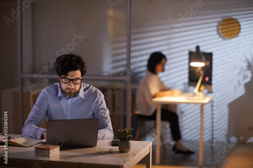 Young bearded businessman in eyeglasses working at his workplace with laptop with businesswoman in the background
