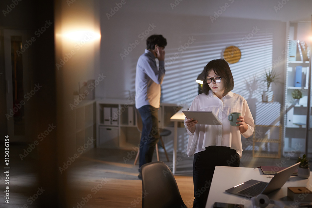 Asian businesswoman looking at tablet pc in her hands and drinking coffee with businessman talking on the phone in the background Asian businesswoman looking at tablet pc in her hands and drinking coffee with businessman talking on the phone in the background