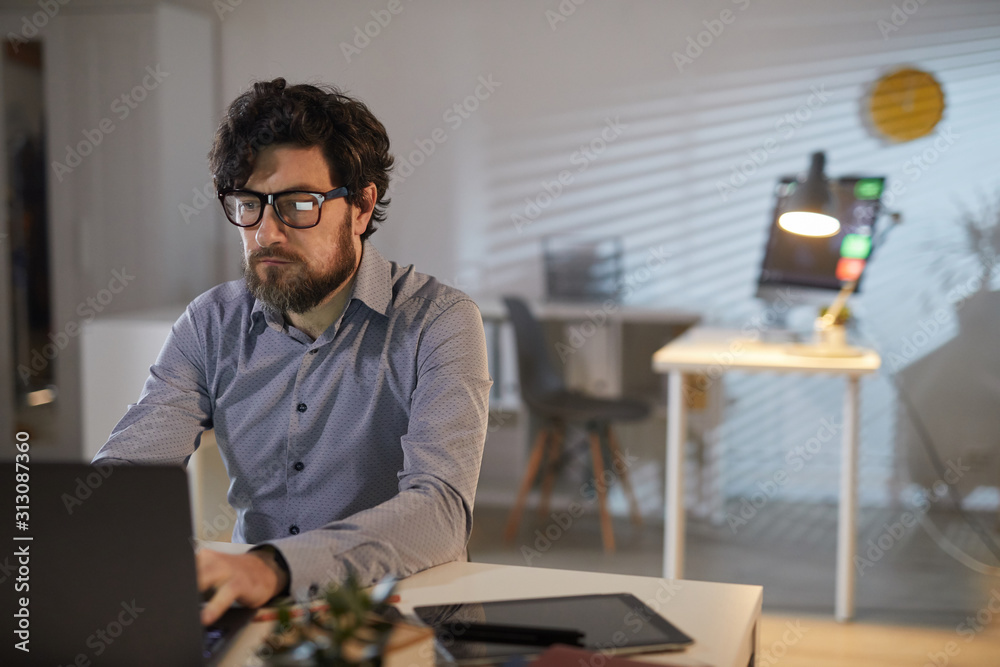 Young bearded designer in eyeglasses sitting at the table and concentrating on his work on laptop at office Young bearded designer in eyeglasses sitting at the table and concentrating on his work on laptop at office