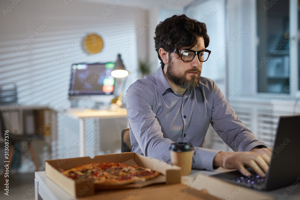 Serious bearded businessman in eyeglasses typing on laptop and eating pizza during his lunch at office Serious bearded businessman in eyeglasses typing on laptop and eating pizza during his lunch at office