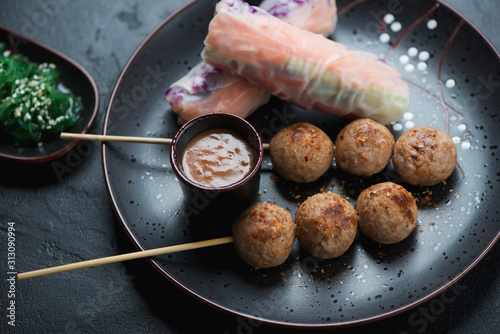 Nem nuong or fried vietnamese meatball skewers with spring rolls and dip sauce served on a black plate, selective focus, closeup photo