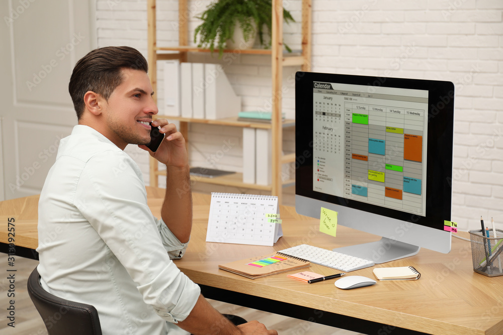 Man talking on smartphone while working with calendar app at table in office Man talking on smartphone while working with calendar app at table in office