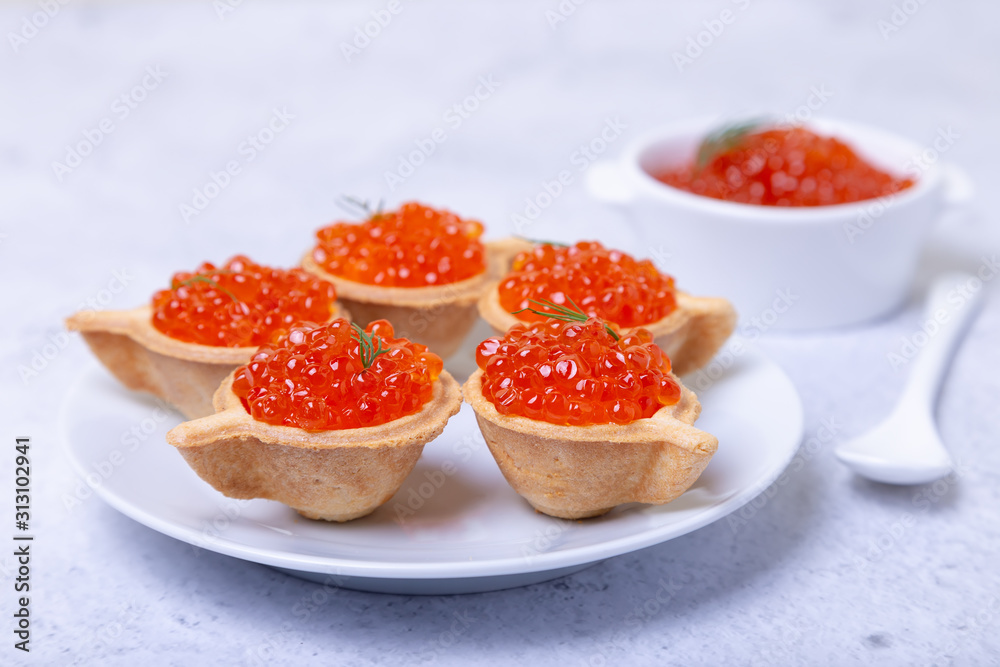 Red caviar (salmon caviar) in tartlets. In the background is a white bowl with caviar. Selective focus, close-up.