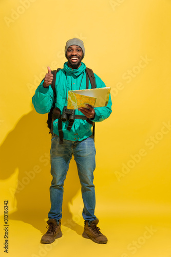 Full length portrait of a cheerful young african-american tourist guy with bag and map isolated on yellow studio background. Preparing for traveling, looking for trip way, smiling and greeting. © master1305