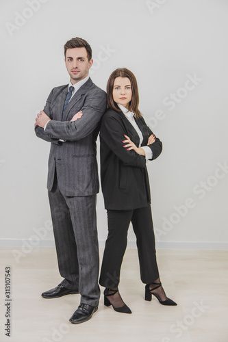 Full-length business man and woman standing alongside, looking confidently at the camera, their hands folded.
