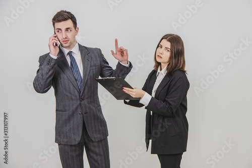 Handsome busy businessman is talking on the phone, pointing his finger up for his secretary to wait. Secretery standing with clipboard, waiting for boss's signature. photo