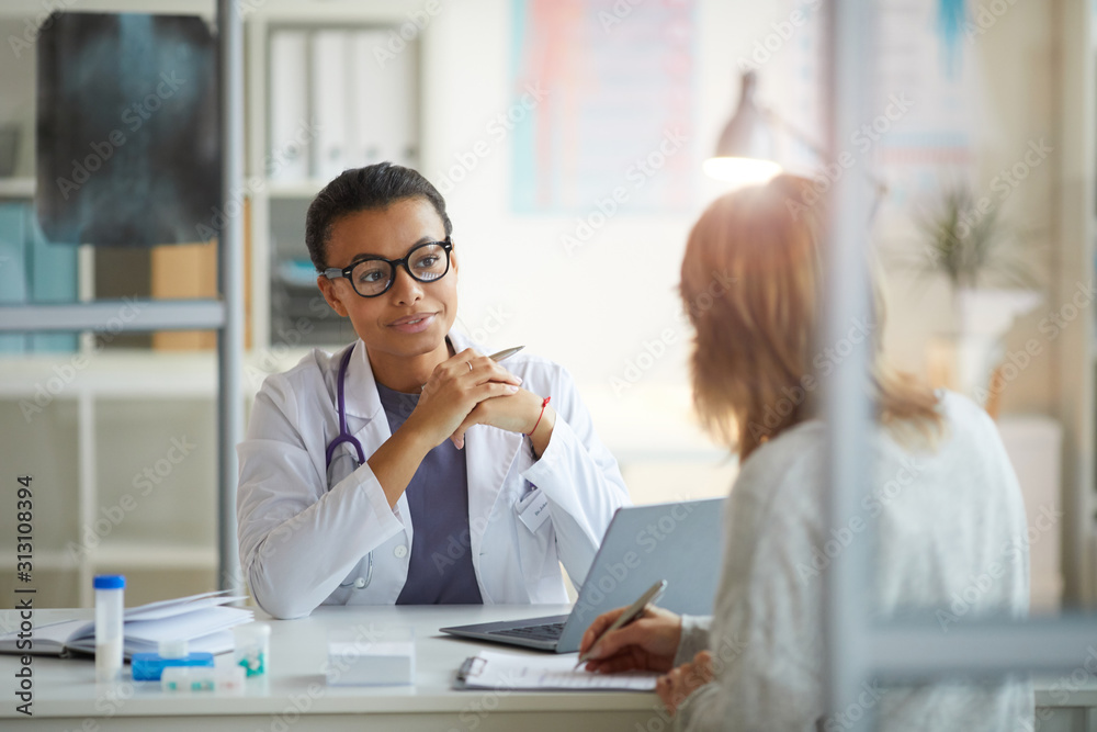 Fototapeta premium Young female doctor in white coat sitting at the table and listening to patient sitting in front of her at office