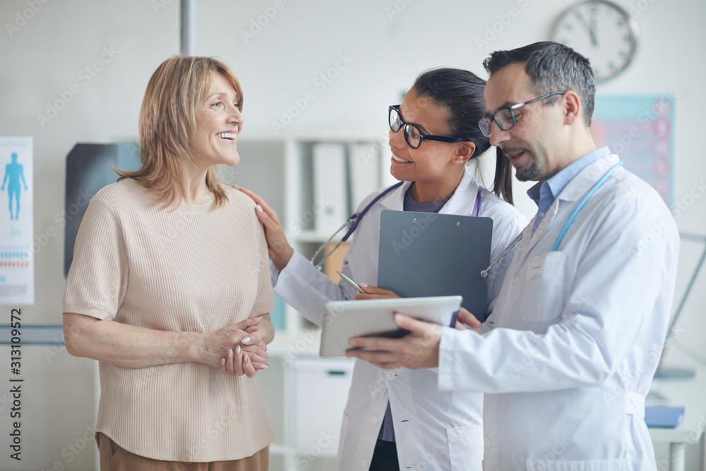 Male doctor and nurse using digital tablet and discussing with smiling mature woman her treatment while they standing at office Male doctor and nurse using digital tablet and discussing with smiling mature woman her treatment while they standing at office