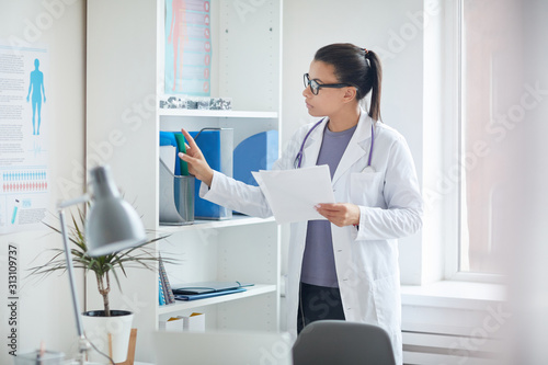 Female doctor in white coat standing near the bookcase and looking for medical cards of her patients at medical clinic