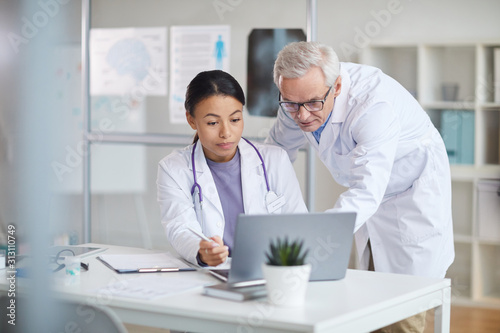 Senior doctor in white coat and young female nurse looking at monitor of laptop and discussing online presentation at office
