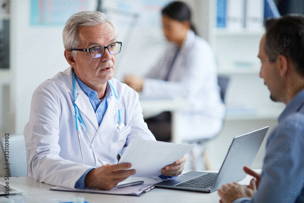 Senior doctor in eyeglasses and in white coat sitting at the table with document and explaining the treatment to his patient at office Senior doctor in eyeglasses and in white coat sitting at the table with document and explaining the treatment to his patient at office