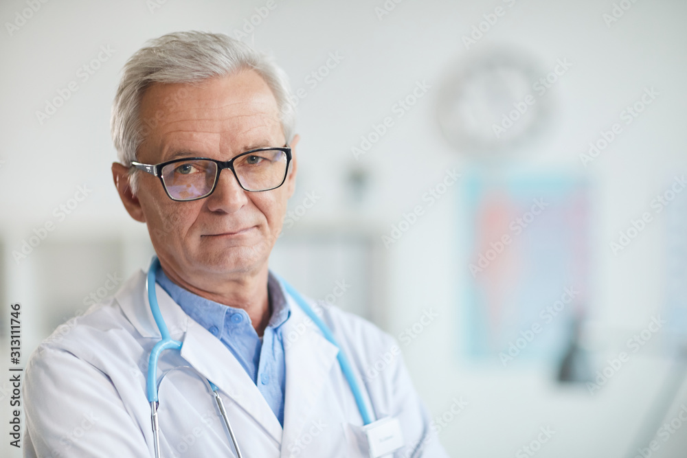 Portrait of experienced senior doctor in eyeglasses and in white coat looking at camera while standing at office Portrait of experienced senior doctor in eyeglasses and in white coat looking at camera while standing at office