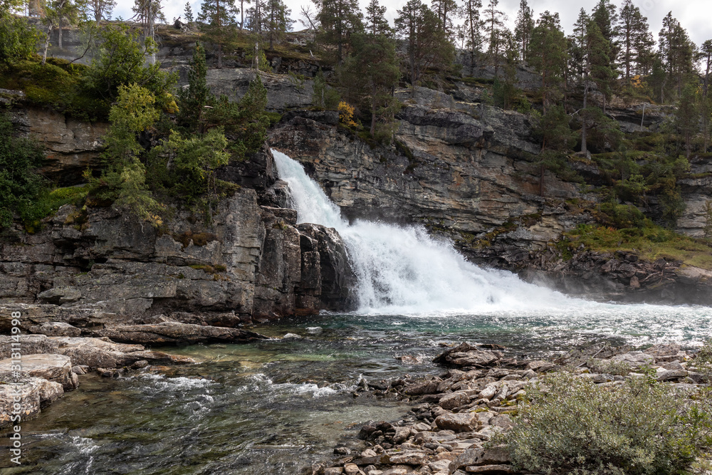 Obraz premium Scenic waterfall on river in mountains of Norway with rocky wall and pine trees, wild nature northern landscape