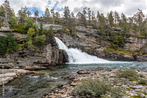 Scenic waterfall on river in mountains of Norway with rocky wall and pine trees, wild nature northern landscape