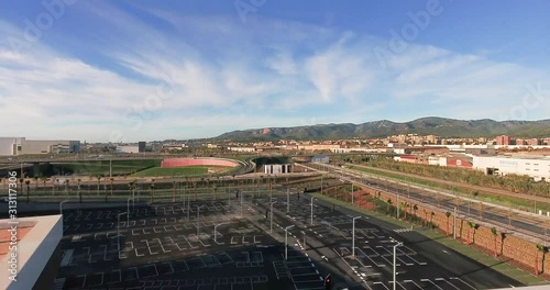 Aerial view over commercial area in the suburbs of Barcelona, Spain, showing a parking with small palm trees, and highway roads, a bridge and soft mountains in this rural background