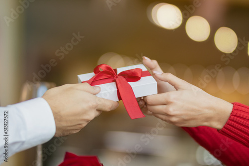 A young woman takes a present giftbox with red ribbon from her boyfriend. Warm and lovely background of a restaurant. Two glasses of wine and a rose on the cafe's table. Valentine's day concept.
