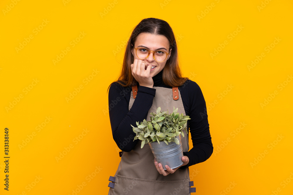 Young gardener girl holding a plant over isolated yellow background nervous and scared Young gardener girl holding a plant over isolated yellow background nervous and scared