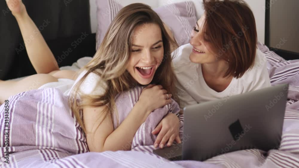 Two joyful laughing lesbians are lying in bed at home and are looking at the laptop screen. Two joyful laughing lesbians are lying in bed at home and are looking at the laptop screen.