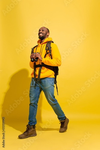 Smiling, looks happy. Portrait of a cheerful young african-american tourist guy with bag and binoculars isolated on yellow studio background. Preparing for traveling. Resort, human emotions, vacation.