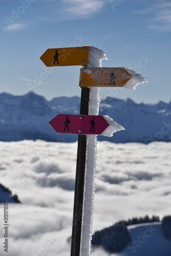 Hiking trail signs with snow and ice on a swiss mountain in winter. Mountains and clouds in the background © Hajnalka