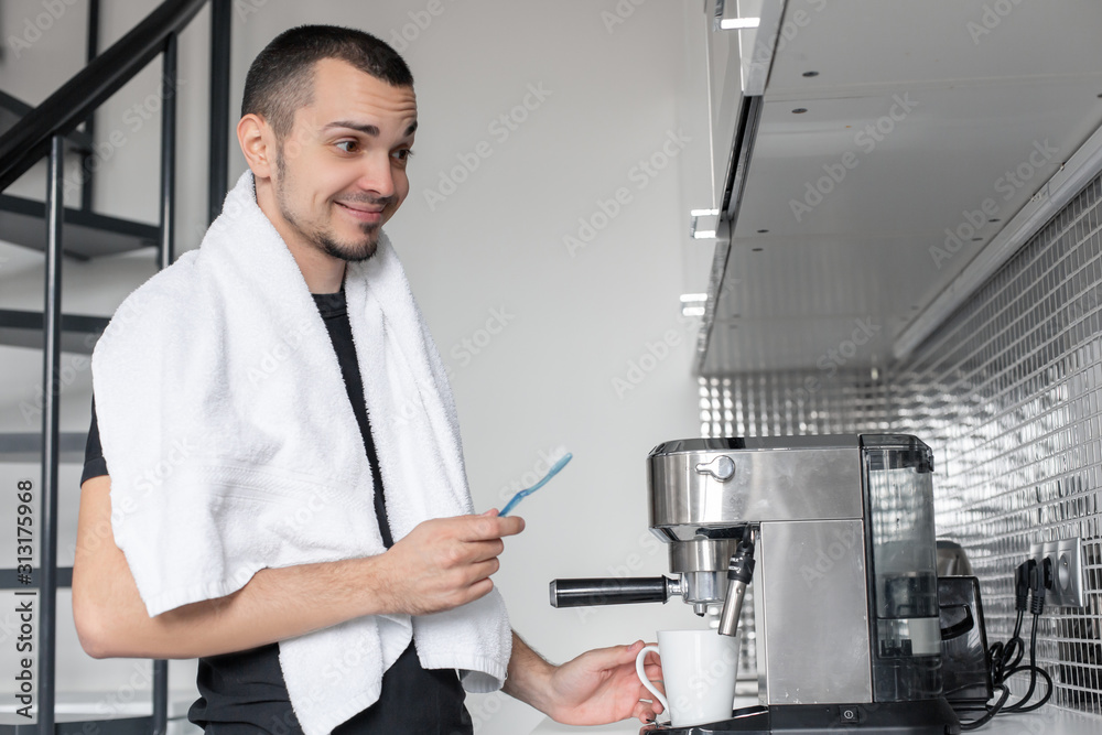 Fototapeta premium A young guy is going to work in the morning. Brushes teeth near a coffee machine while waiting for a cup of coffee