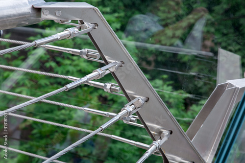 a corner of a glass bridge with tension steel cables and a cable tensioner with thread over green trees.