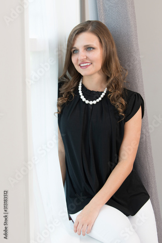 Beautiful young Caucasian woman poses in home wearing stylish black blouse and white pants with pearls - sitting in window looking out