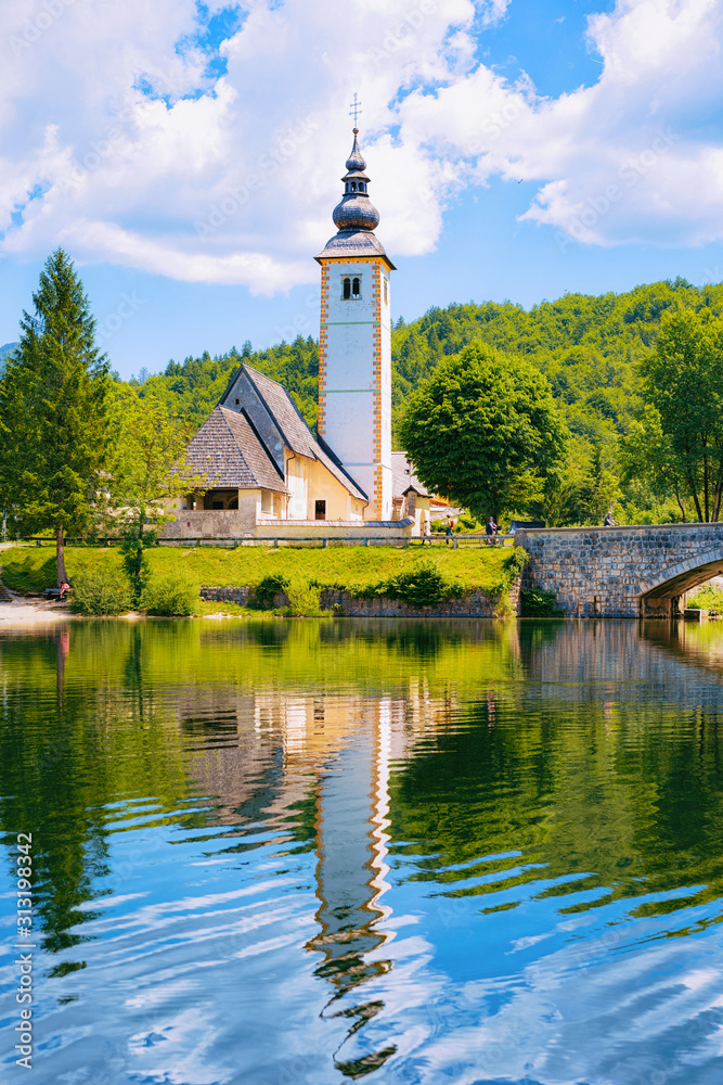 Scenery with Church of St John Baptist on Bohinj Lake in Slovenia. Nature in Slovenija. View of blue sky with clouds. Beautiful landscape in summer. Alpine Travel destination. Julian Alps mountains