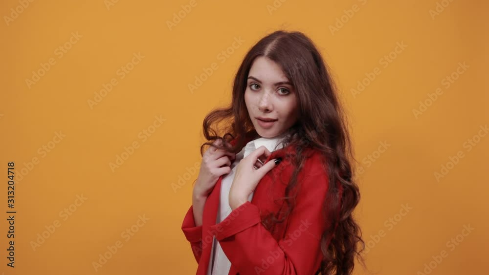 Beautiful brunette young woman in fashion white shirt and red jacket keeping hands on jacket, staying sideways isolated on orange background in studio. People sincere emotions, lifestyle concept. Beautiful brunette young woman in fashion white shirt and red jacket keeping hands on jacket, staying sideways isolated on orange background in studio. People sincere emotions, lifestyle concept.