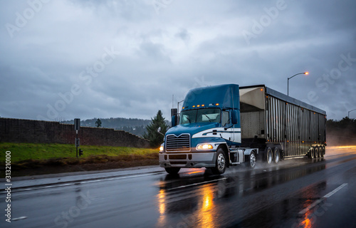 Big rig bonnet blue semi truck transporting cargo in covered bulk semi trailer running on the wet glossy road with raining weather evening