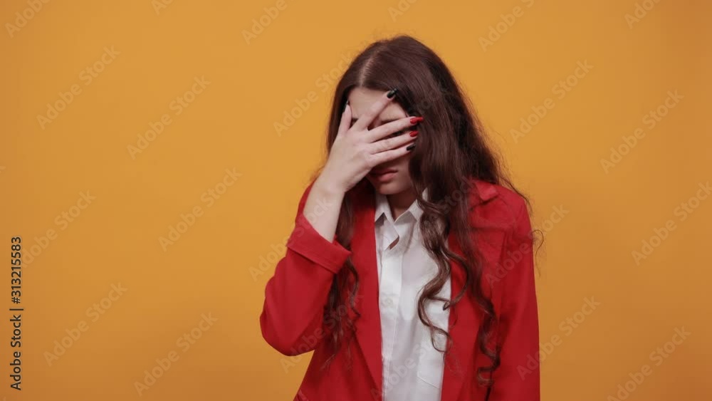 Caucasian young woman in fashion white shirt and red jacket covered eyes with hand, looking through it isolated on orange background in studio. People sincere emotions, lifestyle concept. Caucasian young woman in fashion white shirt and red jacket covered eyes with hand, looking through it isolated on orange background in studio. People sincere emotions, lifestyle concept.