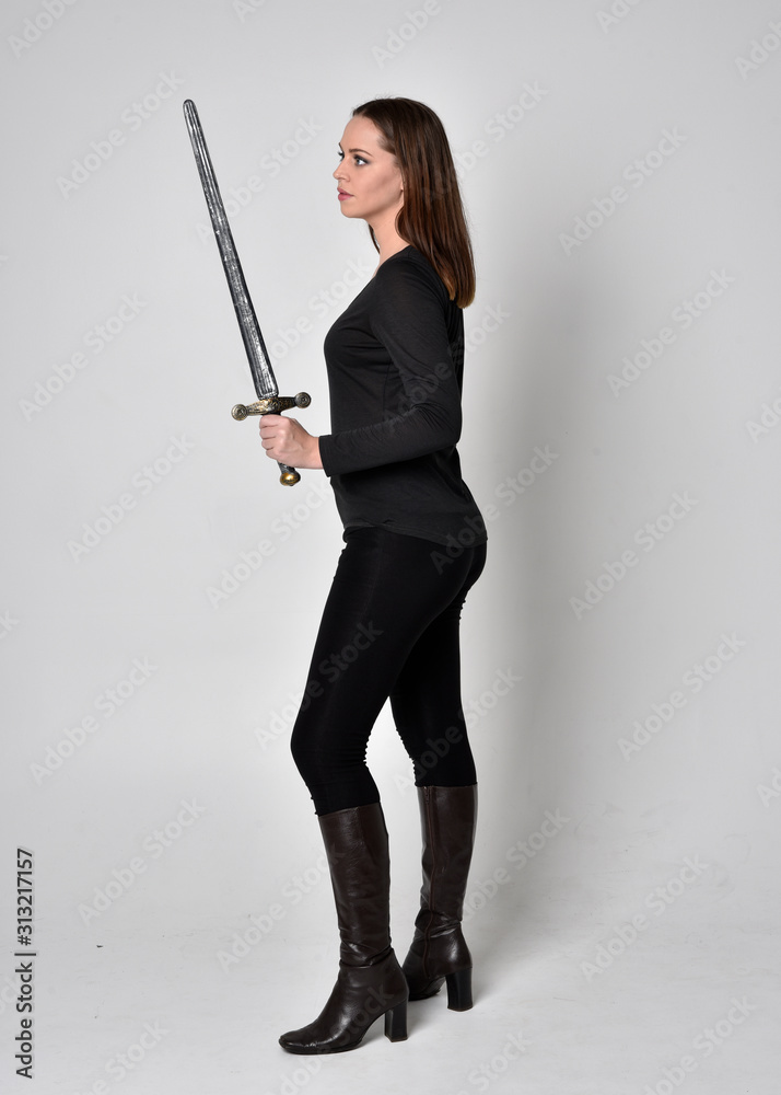 full length portrait of a pretty brunette girl wearing a black shirt and leather boots, holding a sword. Standing pose facing away and holding a sword, on a grey studio background. full length portrait of a pretty brunette girl wearing a black shirt and leather boots, holding a sword. Standing pose facing away and holding a sword, on a grey studio background.
