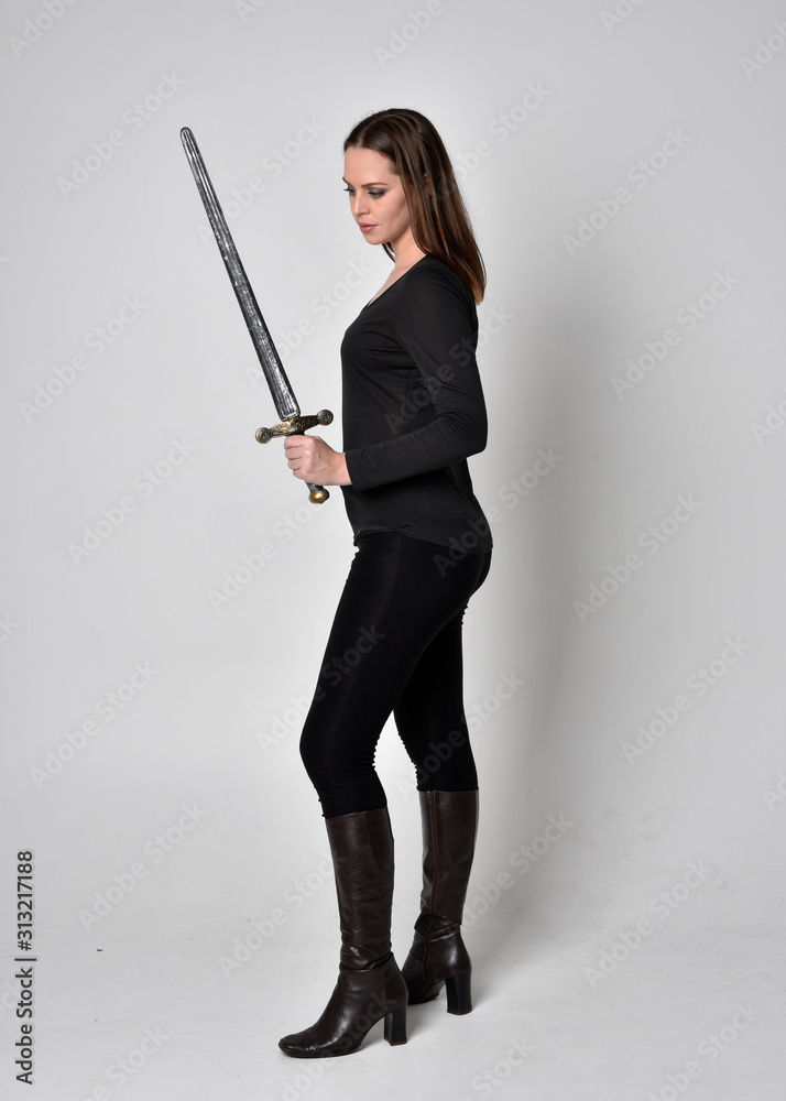 full length portrait of a pretty brunette girl wearing a black shirt and leather boots, holding a sword. Standing pose facing away and holding a sword, on a grey studio background. full length portrait of a pretty brunette girl wearing a black shirt and leather boots, holding a sword. Standing pose facing away and holding a sword, on a grey studio background.