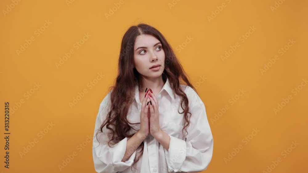 Serious brunette young woman in fashion white shirt keeping hinds together, having plan isolated on orange background in studio. People sincere emotions, lifestyle concept.