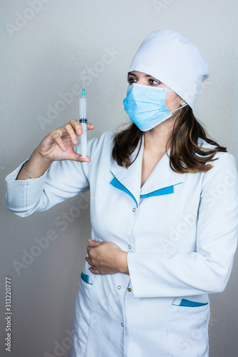 doctor woman in white coat with dark hair in medical blue mask holds syringe in hands on light background photo