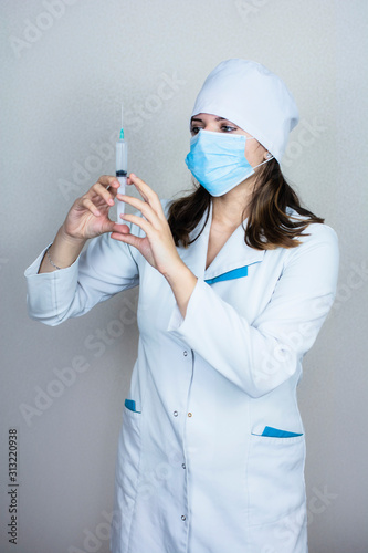 doctor woman in white coat with dark hair in medical blue mask holds syringe in hands on light background photo