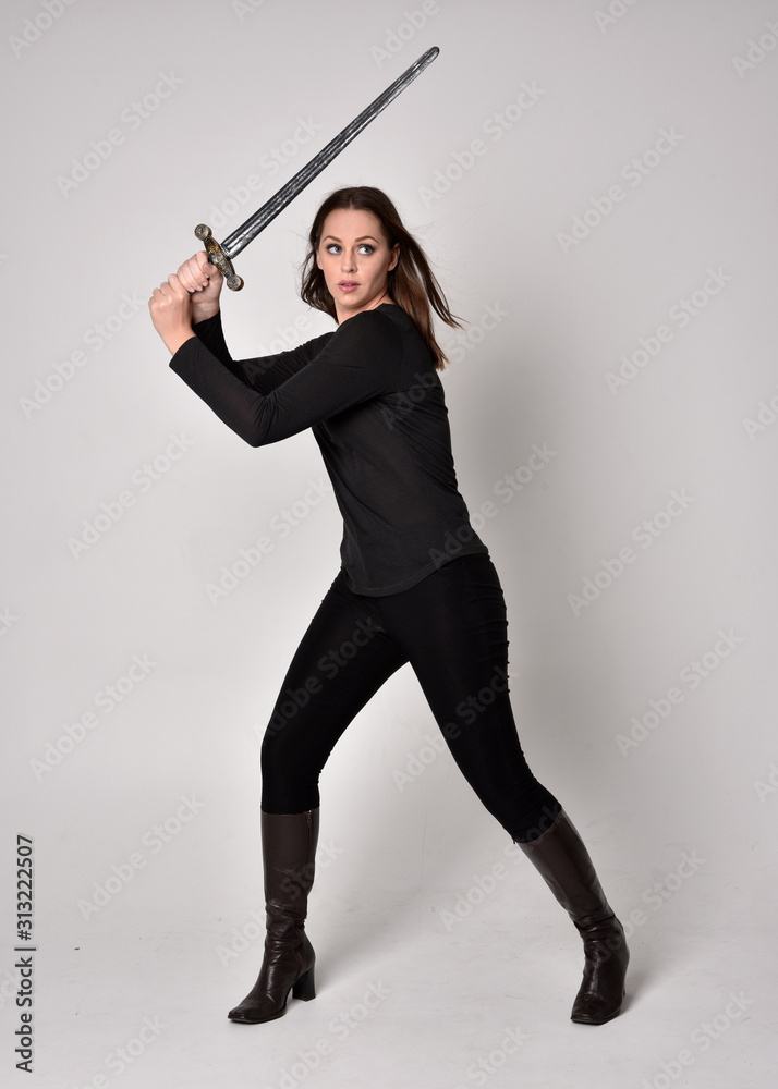 full length portrait of a pretty brunette girl wearing a black shirt and leather boots, holding a sword. Standing pose, holding a sword, on a grey studio background. full length portrait of a pretty brunette girl wearing a black shirt and leather boots, holding a sword. Standing pose, holding a sword, on a grey studio background.