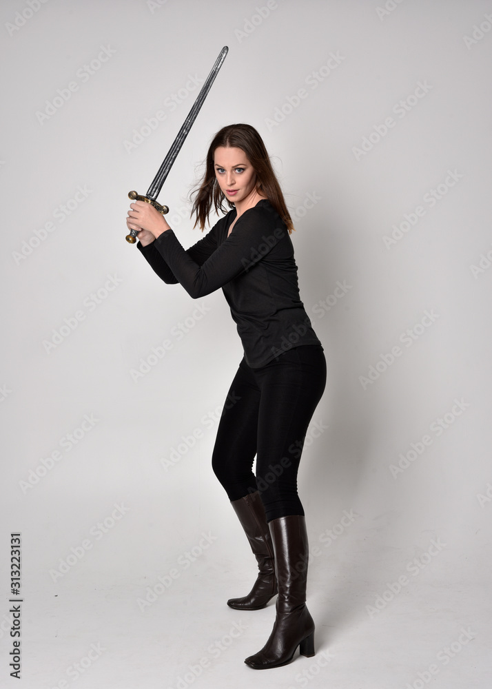 full length portrait of a pretty brunette girl wearing a black shirt and leather boots, holding a sword. Standing pose, holding a sword, on a grey studio background. full length portrait of a pretty brunette girl wearing a black shirt and leather boots, holding a sword. Standing pose, holding a sword, on a grey studio background.