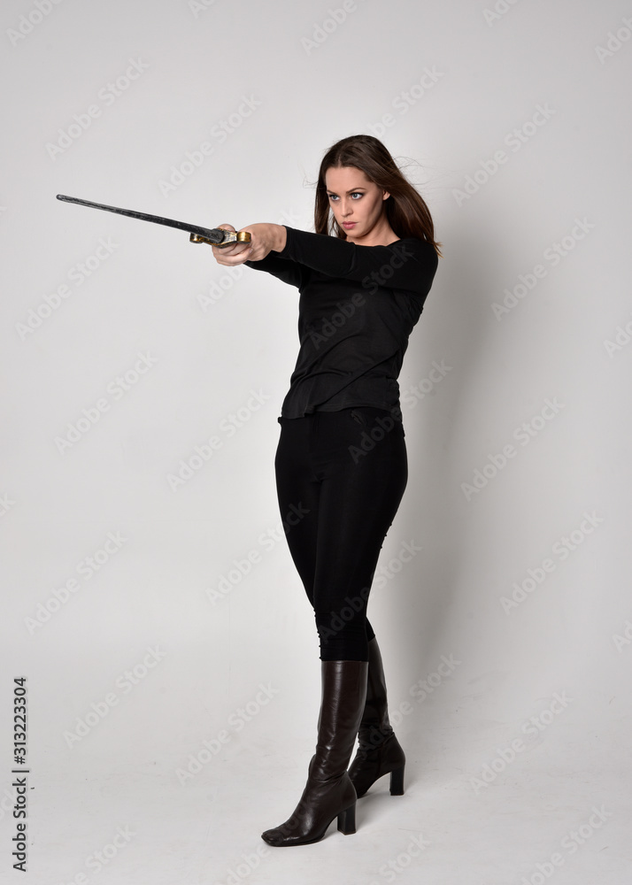 full length portrait of a pretty brunette girl wearing a black shirt and leather boots, holding a sword. Standing pose, holding a sword, on a grey studio background. full length portrait of a pretty brunette girl wearing a black shirt and leather boots, holding a sword. Standing pose, holding a sword, on a grey studio background.