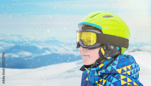 Portrait of a little boy in skier suit, halmet and glasses on ski resort mountain peak. Copy space beside