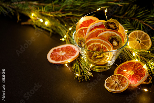 Creative moody holiday Christmas New Year food fruits with dried grapefruit, kiwi, orange and lemon in glass bowl with branch of fir tree with warm led lights, angle view, copy space
