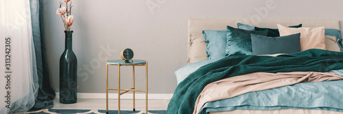Flowers in bottle shape vase next to clock on stylish nightstand with golden legs next to king size bed with emerald green, blue and beige bedding