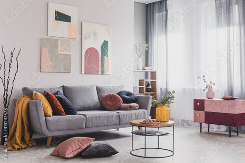 Real photo of abstract paintings hanging on white wall above a gray sofa in a living room interior with big windows photo