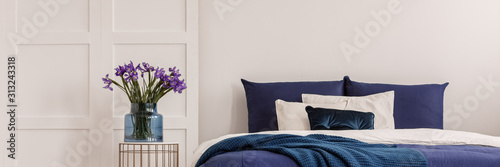 Purple flowers in blue glass vase on stylish bedside table next to king size bed