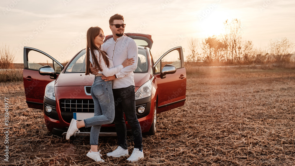 Young Happy Couple Dressed Alike in White Shirt and Jeans Enjoying Road Trip at Their New Car, Beautiful Sunset on the Field, Vacation and Travel Concept Young Happy Couple Dressed Alike in White Shirt and Jeans Enjoying Road Trip at Their New Car, Beautiful Sunset on the Field, Vacation and Travel Concept