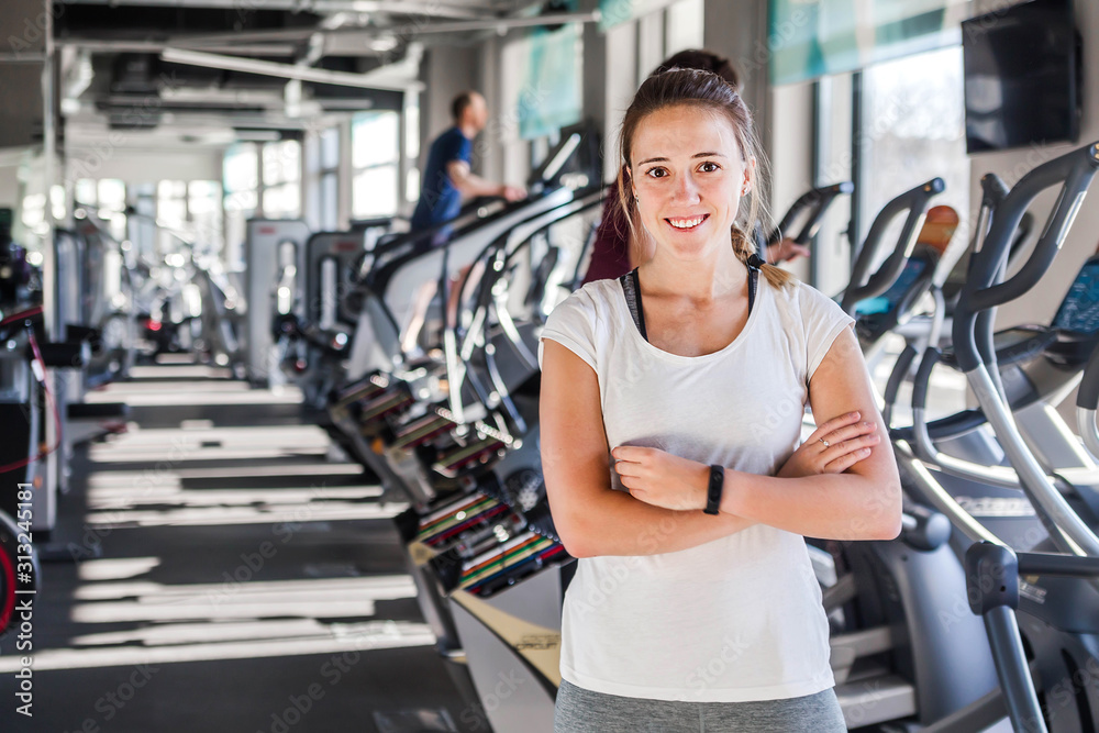 Smiling sports caucasian woman standing with arms cross and looking camera at fitness gym background with copy space in left. Trainer or instructor fitness coach concept.....C Smiling sports caucasian woman standing with arms cross and looking camera at fitness gym background with copy space in left. Trainer or instructor fitness coach concept.....C