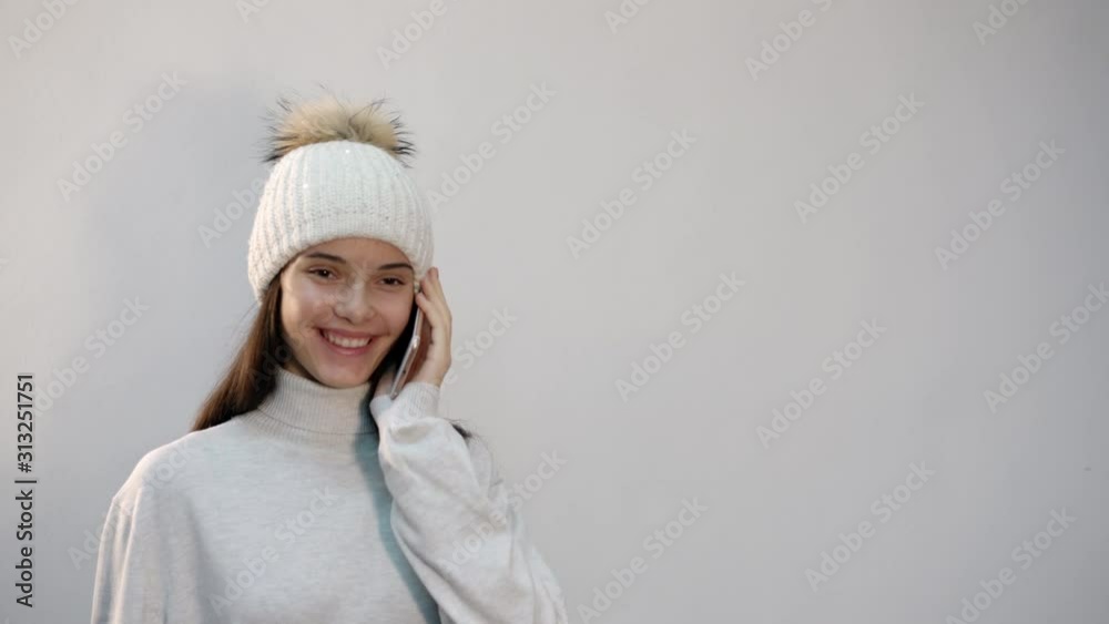 happy pretty girl teenager in white warm hat and sweater talks on mobile phone on light background in studio happy pretty girl teenager in white warm hat and sweater talks on mobile phone on light background in studio