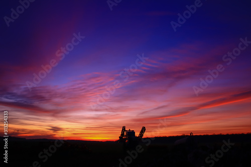 Incredible very colorful sunset in the countryside. Silhouette of an industrial agricultural machine against the day. Cumulonimbus and stratus in a beautiful sky at night.